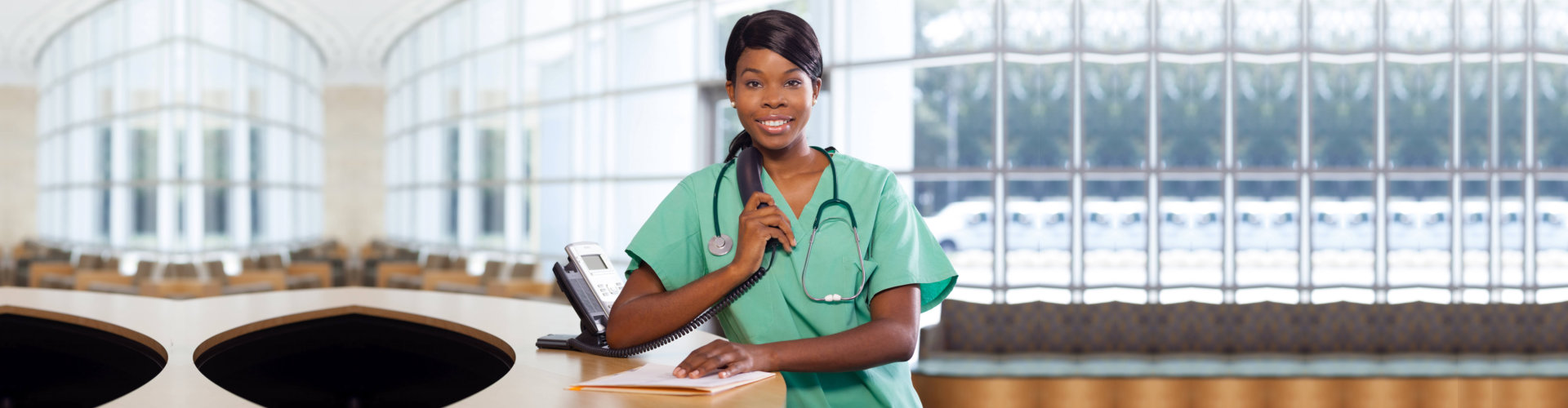 Smiling African American nurse at hospital work station lit brightly with phone and stethoscope.